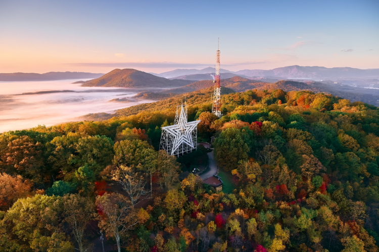 An aerial view of a mountainous landscape during sunrise, showcasing a star-shaped structure and a communication tower surrounded by vibrant autumn foliage. A misty valley is visible in the background, enhancing the tranquil atmosphere of the scene.