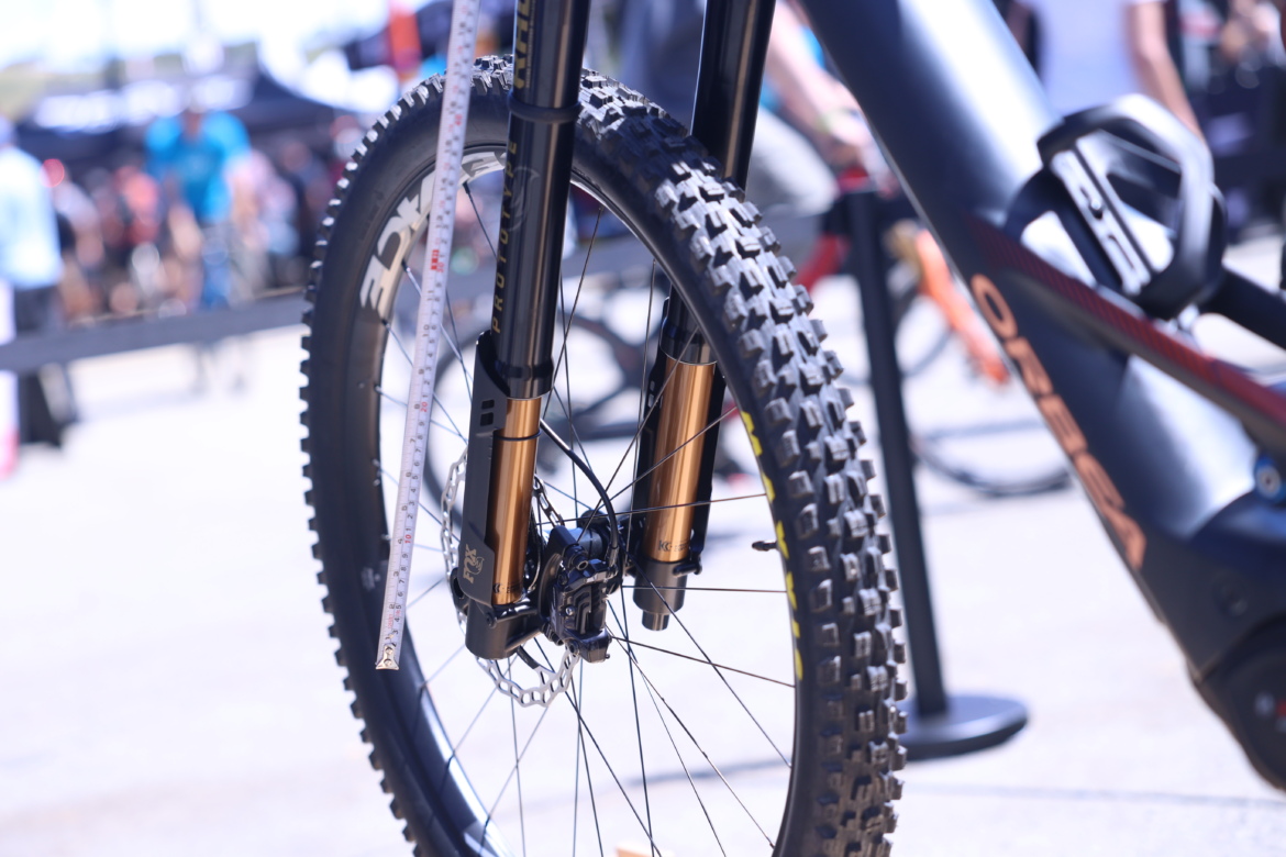 Close-up view of a mountain bike front wheel, highlighting the suspension forks with gold accents and a black tire featuring a knobby tread pattern. A measuring tape is positioned alongside the forks, indicating attention to bike setup and maintenance. In the background, people are blurred, suggesting a lively outdoor event or trade show.