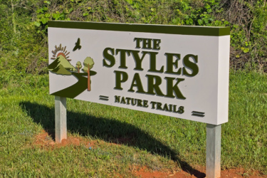 Sign for "The Styles Park" featuring the words "Nature Trails," with graphic elements including mountains, trees, and a bird, set against a green landscape.