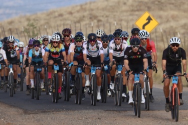 A large group of cyclists participating in a race, riding along a rural road with a slight incline. They are wearing colorful cycling jerseys and helmets, with various race numbers visible on their bikes. In the background, there are dry hills and a warning sign indicating a curve ahead. Chino Grinder - Gravel Race - Arizona - October 18th, 2025 mountain bike trail.