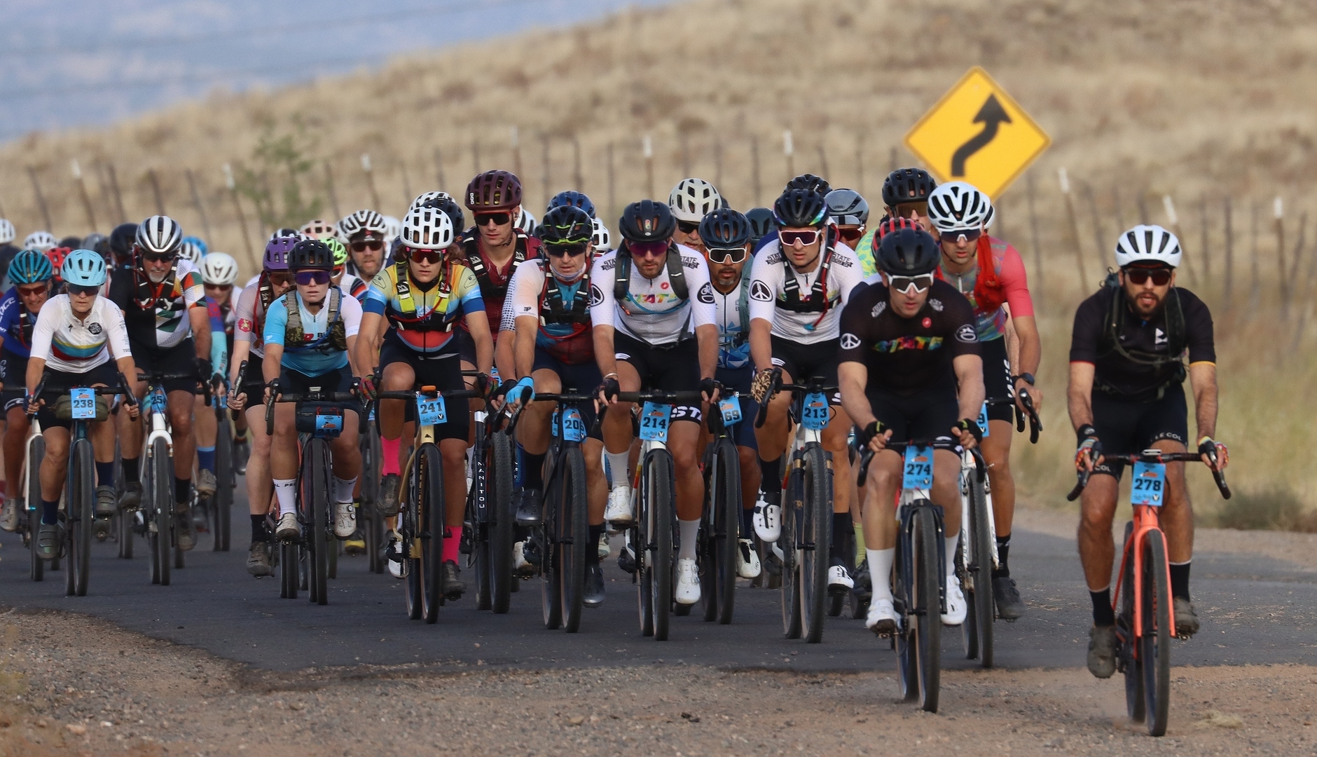 A large group of cyclists participating in a race, riding along a rural road with a slight incline. They are wearing colorful cycling jerseys and helmets, with various race numbers visible on their bikes. In the background, there are dry hills and a warning sign indicating a curve ahead. Chino Grinder - Gravel Race - Arizona - October 18th, 2025 mountain bike trail.