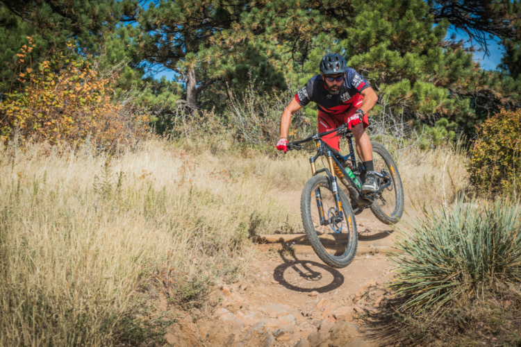 A mountain biker performing a jump on a dirt trail surrounded by tall grass and trees, with a clear blue sky in the background. The rider is wearing a helmet and padded gear for safety.
