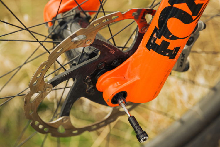 Close-up view of a mountain bike's front fork, featuring an orange FOX logo and a visible disc brake rotor, set against a blurred background of grass.