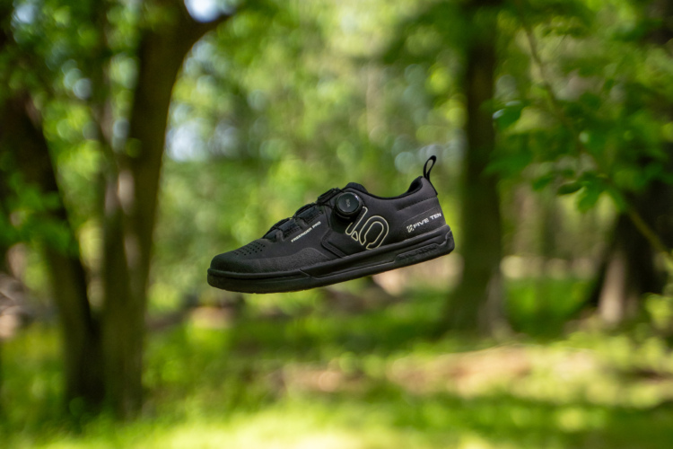 A black athletic shoe designed for outdoor activities, suspended in mid-air against a blurred green forest background. The shoe features a sleek design with a snug fit, a dial for securing the laces, and includes branding on the side.