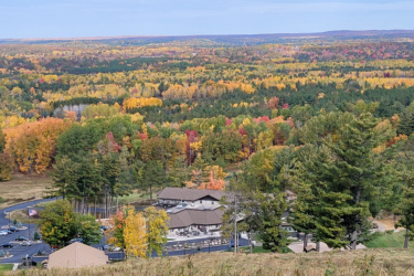 A panoramic view of a colorful forest during autumn, showcasing vibrant shades of orange, yellow, and red. In the foreground, a building and parking area are visible, surrounded by lush greenery. The landscape extends into the distance, featuring a varied tree line and rolling hills under a clear blue sky. Pine Mountain Resort mountain bike trail.