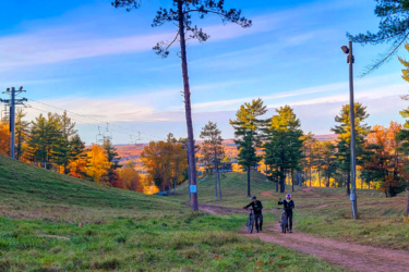 Two cyclists walk their bikes along a dirt path on a hillside surrounded by colorful autumn trees. In the background, a ski lift and a ski jump structure are visible under a clear blue sky. The scene captures the beauty of fall and outdoor recreation. Pine Mountain Resort mountain bike trail.