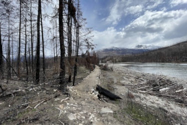 A path runs alongside a river in a landscape affected by wildfire. Scorched black trees stand in the background, contrasting with the mountainous terrain and cloudy sky. The foreground features a rocky riverbank with fallen logs, highlighting the impact of the fire on the surrounding environment. 12 to 5 to 5 / Valley of the Five Lakes and Wabasso Lake mountain bike trail.