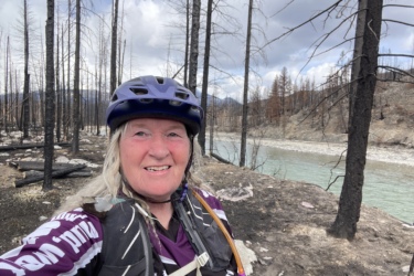 A smiling woman with long hair is standing near a river, wearing a purple helmet and a cycling jersey. The background depicts a landscape affected by a wildfire, featuring charred trees and a rocky terrain. The sky is partly cloudy, and the river appears calm. 12 to 5 to 5 / Valley of the Five Lakes and Wabasso Lake mountain bike trail.