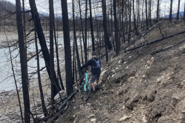 A person standing next to a bicycle on a dirt path next to a river, surrounded by tall, charred trees in a post-wildfire landscape. The sky is partly cloudy, and the ground is rocky and uneven. 12 to 5 to 5 / Valley of the Five Lakes and Wabasso Lake mountain bike trail.