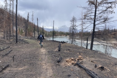 A person riding a mountain bike along a trail in a forest affected by wildfire, with burned trees on either side. In the background, a river and snow-capped mountains can be seen under a cloudy sky. 12 to 5 to 5 / Valley of the Five Lakes and Wabasso Lake mountain bike trail.