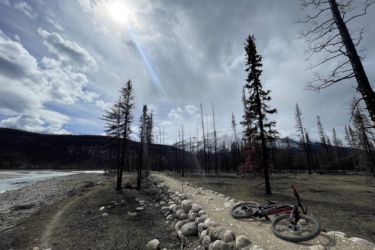 A mountain bike resting on a gravel path alongside a river, surrounded by a landscape of tall, barren trees against a backdrop of mountains and a cloudy sky. The sun peeks through the clouds, illuminating the scene. 12 to 5 to 5 / Valley of the Five Lakes and Wabasso Lake mountain bike trail.