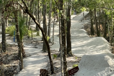 A winding dirt path through a forested area, surrounded by dense trees and greenery. The path is light-colored and appears well-maintained, with a sign indicating "One Way - Do Not Enter." In the background, a person is seen standing on the path. Alafia River State Park mountain bike trail.