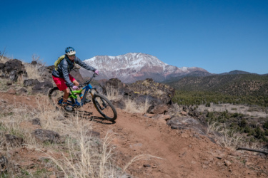 A mountain biker is navigating a dirt trail on a rocky landscape, with a snow-capped mountain visible in the background under a clear blue sky. The rider is wearing a helmet, a long-sleeve shirt, and shorts, showcasing a dynamic riding position as they lean into the turn. Cliffrose Trail System mountain bike trail.