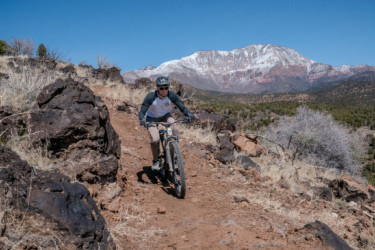 A mountain biker navigating a rocky trail with a backdrop of snow-capped mountains and a clear blue sky. The terrain features dry grass and scattered rocks, indicating a natural outdoor setting. Cliffrose Trail System mountain bike trail.