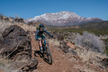 A mountain biker navigates a rocky trail with a mountainous background under a clear blue sky. Snow-capped peaks and diverse vegetation enhance the scenic landscape, showcasing the thrill of outdoor adventure. Cliffrose Trail System mountain bike trail.