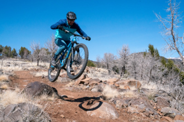A mountain biker in a blue and teal outfit jumps high over a rocky trail, showcasing an action-packed moment in a natural landscape with dry vegetation and a clear blue sky. Cliffrose Trail System mountain bike trail.