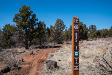 Wooden trail marker indicating the "PJ Party" trail, with symbols for bike access, difficulty level as "easiest," and a sign stating "closed to motor vehicles." Surrounding landscape features sparse vegetation and blue sky in the background, with a dirt path leading into the distance.