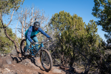 A mountain biker navigates rocky terrain surrounded by trees on a clear day, wearing a helmet and colorful cycling gear. Cliffrose Trail System mountain bike trail.