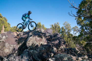 A mountain biker riding over large rocks in a natural outdoor setting, with trees and a clear blue sky in the background. The cyclist is wearing a helmet and a blue and teal outfit.