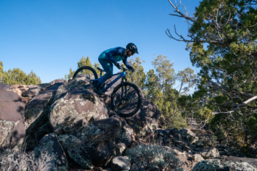 A mountain biker skillfully navigates a rocky terrain, perched on a large boulder, with trees and a clear blue sky in the background. The biker is wearing a helmet and a blue outfit, demonstrating agility and control as they ride. Cliffrose Trail System mountain bike trail.