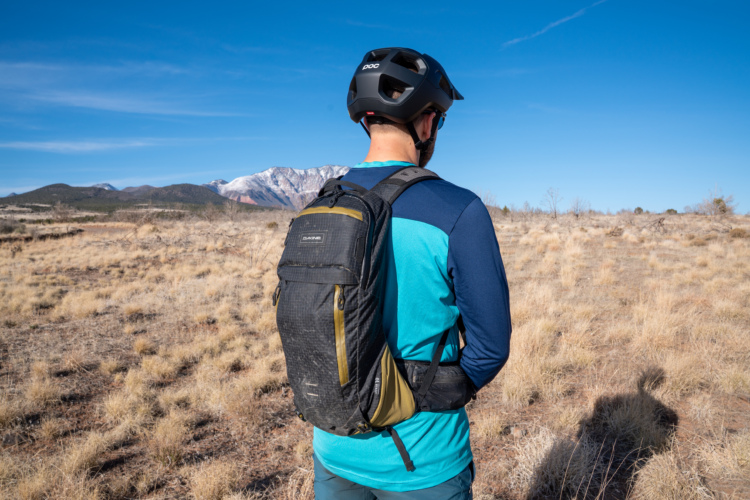 A person wearing a blue and black long-sleeve shirt and a helmet stands in a grassy field, facing away from the camera. They are carrying a black and green backpack. In the background, mountains with snow-capped peaks are visible under a clear blue sky.