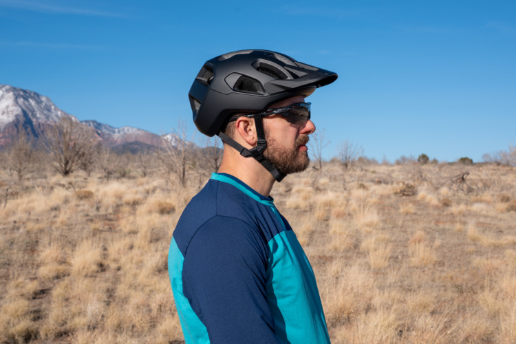 A man wearing a black bicycle helmet and sunglasses stands in profile against a backdrop of mountains and a clear blue sky. He is dressed in a blue and teal athletic shirt, with sparse vegetation and dry grass visible in the foreground.