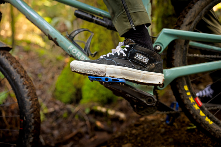 A close-up view of a mountain bike pedal and shoe. A black sneaker with white laces and a textured sole rests on a blue pedal, surrounded by a blurred natural background of greenery and dirt. The bike frame is partially visible, showcasing its design with the word "FORB" on it.