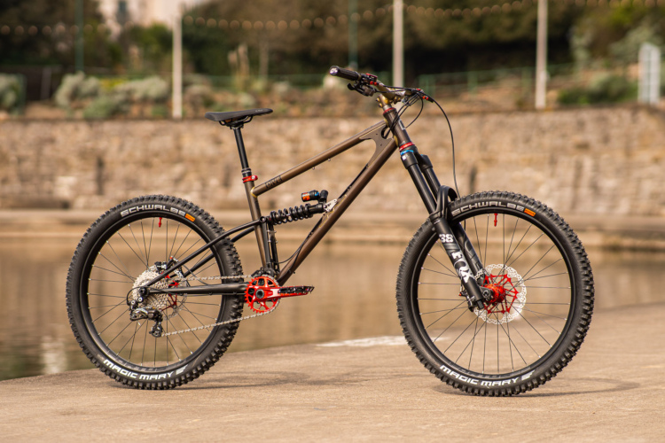 A high-end mountain bike with a sleek, dark frame and vibrant red components parked beside a body of water. The bike features wide tires designed for off-road terrain, front suspension forks, and a prominent rear shock, emphasizing its performance capabilities. The background includes a stone wall and greenery, indicating an outdoor setting.