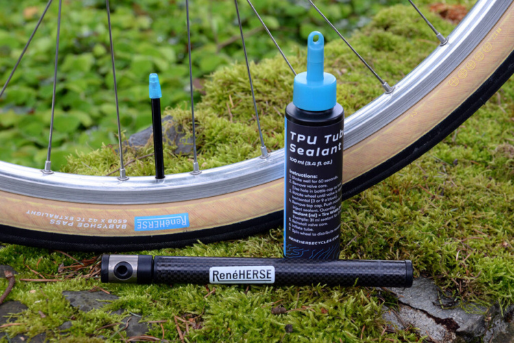 A close-up image of a bicycle wheel showing a tire with a black valve stem, next to a bottle of TPU Tube Sealant and a carbon fiber tire lever. The background features green moss and small plants, highlighting a natural outdoor setting.