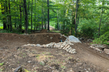 A dirt trail winding through a wooded area, featuring a jump constructed with rocks and dirt. Lush green trees surround the path, creating a serene natural setting. A bicycle is positioned near the jump, suggesting it's designed for mountain biking. Pine Mountain Resort mountain bike trail.