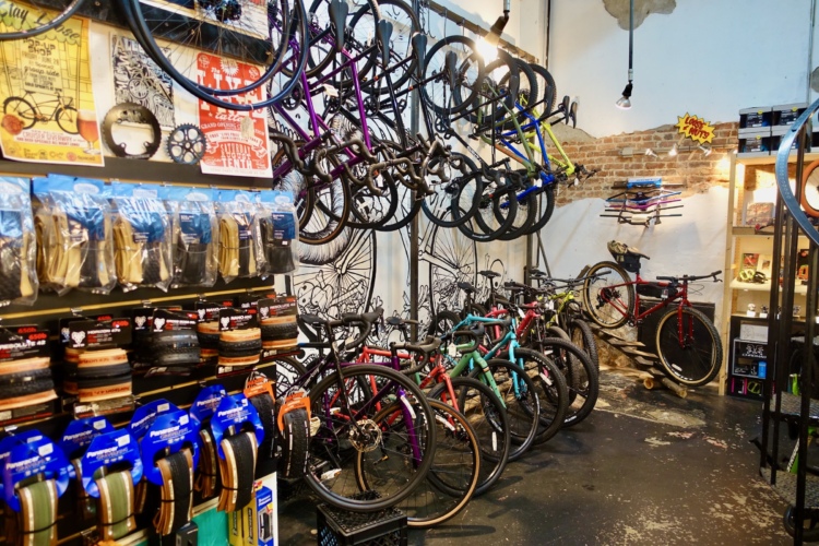 A colorful bicycle shop interior featuring a variety of bicycles on display, including road and mountain bikes. The walls are adorned with bicycle-themed posters, and shelves showcase bike accessories such as tires and grips. The ambiance is vibrant, with a mix of urban art and organized merchandise.