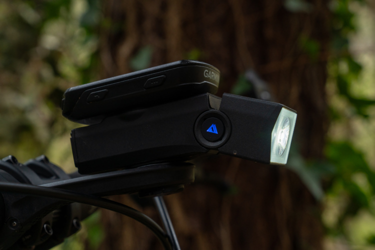 Close-up of a bicycle handlebar attachment featuring a light with a visible LED beam and a GPS device mounted above it, set against a blurred green background of foliage.