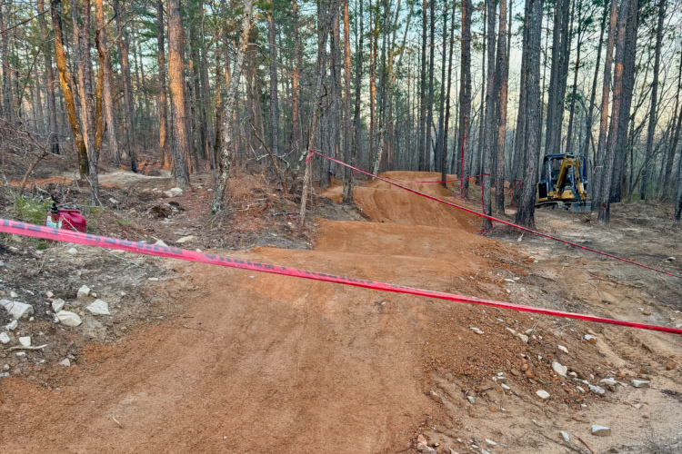 A dirt bike trail under construction in a forest setting, with freshly built sections of the trail visible. Red caution tape is placed across parts of the path, and a piece of construction equipment is seen in the background. The area is surrounded by tall trees and scattered rocks on the ground.
