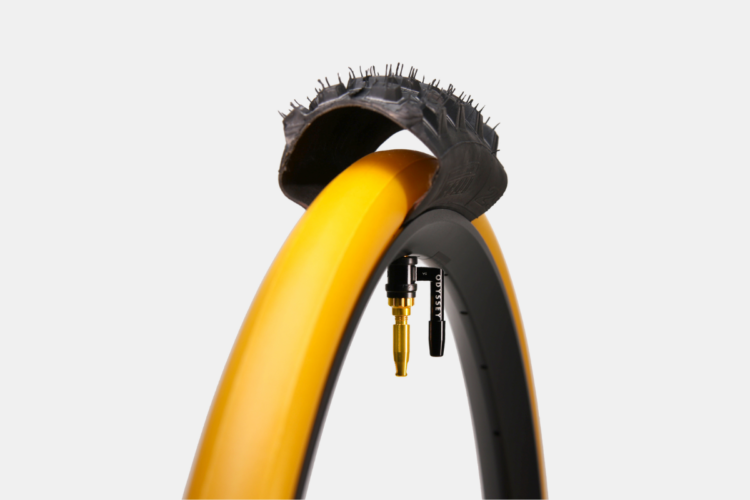 A close-up image of a bicycle tire featuring a black tread with spikes, arching over a bright yellow rim. The tire is mounted on a wheel with a visible valve for inflation, emphasizing its unique design and color contrast. The background is plain white.
