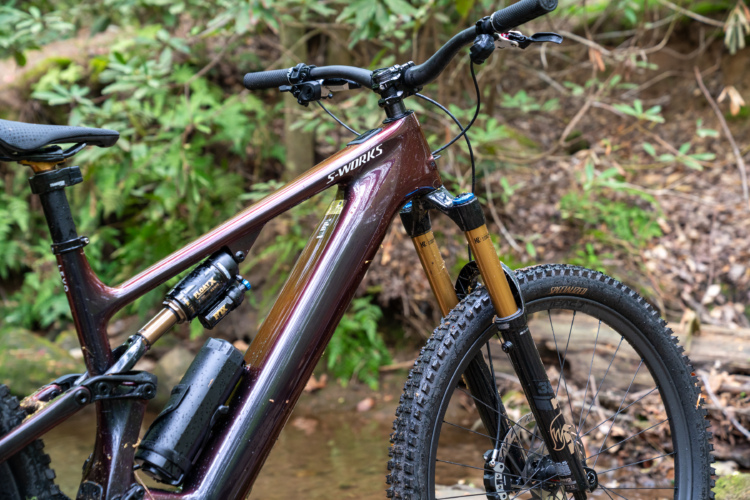 Close-up of a specialized mountain bike frame with a glossy burgundy finish, featuring a suspension system with visible shock absorbers. The handlebars are equipped with grips and brake levers, and the bike has knobby tires. The background contains greenery and a natural setting, suggesting an outdoor environment.