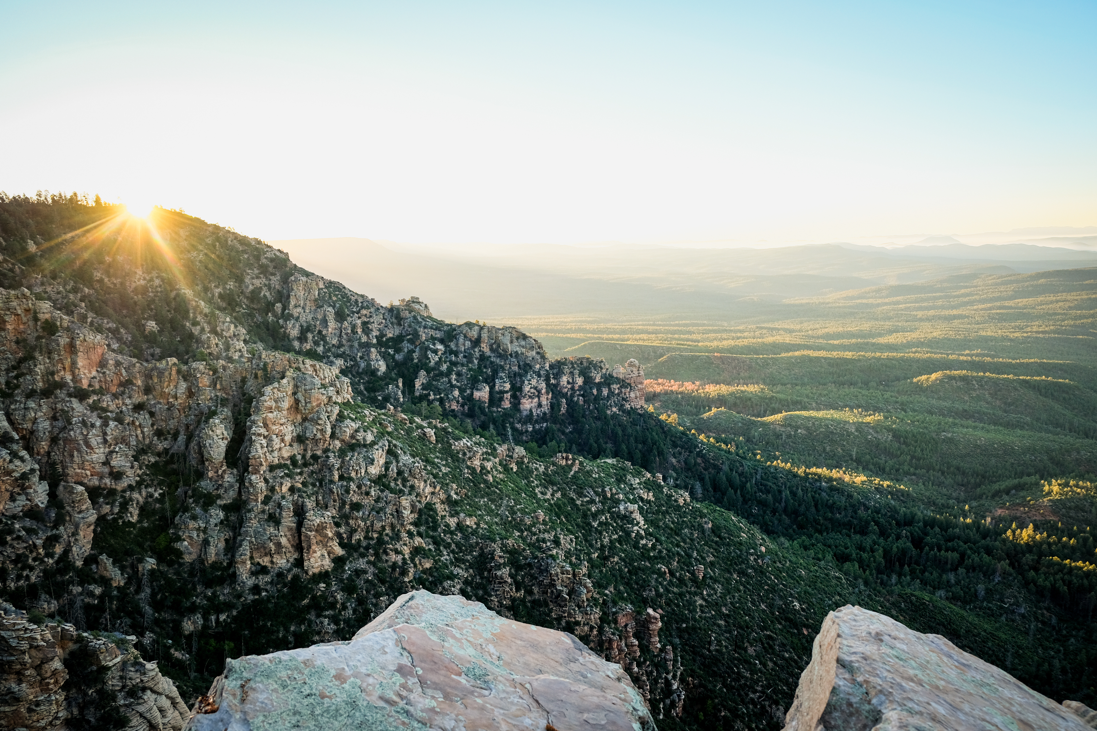 A scenic view of a mountainous landscape at sunrise, featuring rocky cliffs in the foreground and lush green valleys stretching into the distance. The sun casts a warm glow, illuminating the peaks and creating rays of light against a clear sky. Highline Trail mountain bike trail.