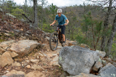 A mountain biker navigating a rocky trail in a forested area, wearing a helmet and athletic gear, with trees in the background and cloudy skies above. Talladega mountain bike trail.