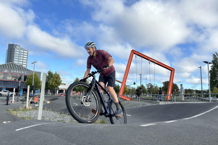 A man riding a bicycle on a pump track in an urban park. He is wearing a helmet and a patterned shirt, smiling as he navigates the course. In the background, there is a modern building labeled "URBANZ" and several people engaging with a play area featuring swings. The sky is partly cloudy with a blue backdrop.