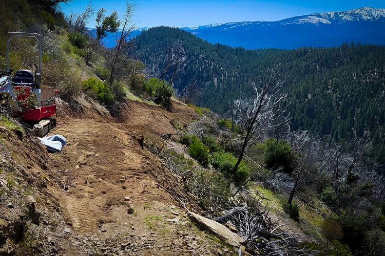A construction site on a hillside, featuring a small piece of machinery parked on a cleared dirt path surrounded by greenery and mountainous terrain. In the background, trees and distant mountains are visible under a clear blue sky.