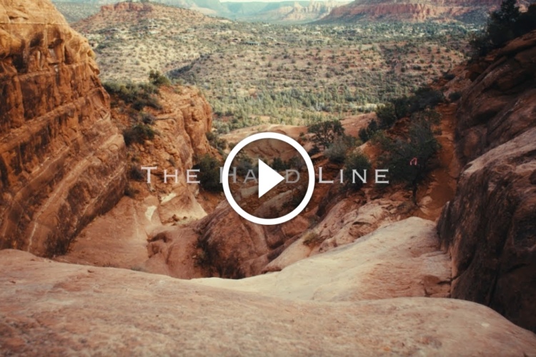 A panoramic view of rugged, desert terrain featuring rocky formations and sparse vegetation, with a scenic valley stretching into the distance. The words "The Hard Line" are superimposed on the image, along with a play button symbol in the center, suggesting that the image may be part of a video or promotional content.