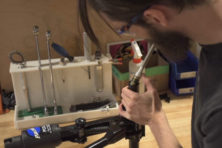 A person with glasses is working on a mechanical assembly, using a tool to assemble a part. Various tools, including wrenches and other mechanical items, are arranged on a workbench in the background.