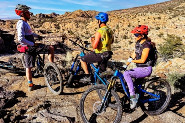 Three mountain bikers in helmets and protective gear are paused on rocky terrain, discussing their ride. They are surrounded by a rugged landscape of hills and sparse vegetation. One rider is on a blue bike, while the others are on black and blue bikes, respectively. The scene captures the spirit of outdoor adventure and camaraderie among cyclists. Granite Dells mountain bike trail.
