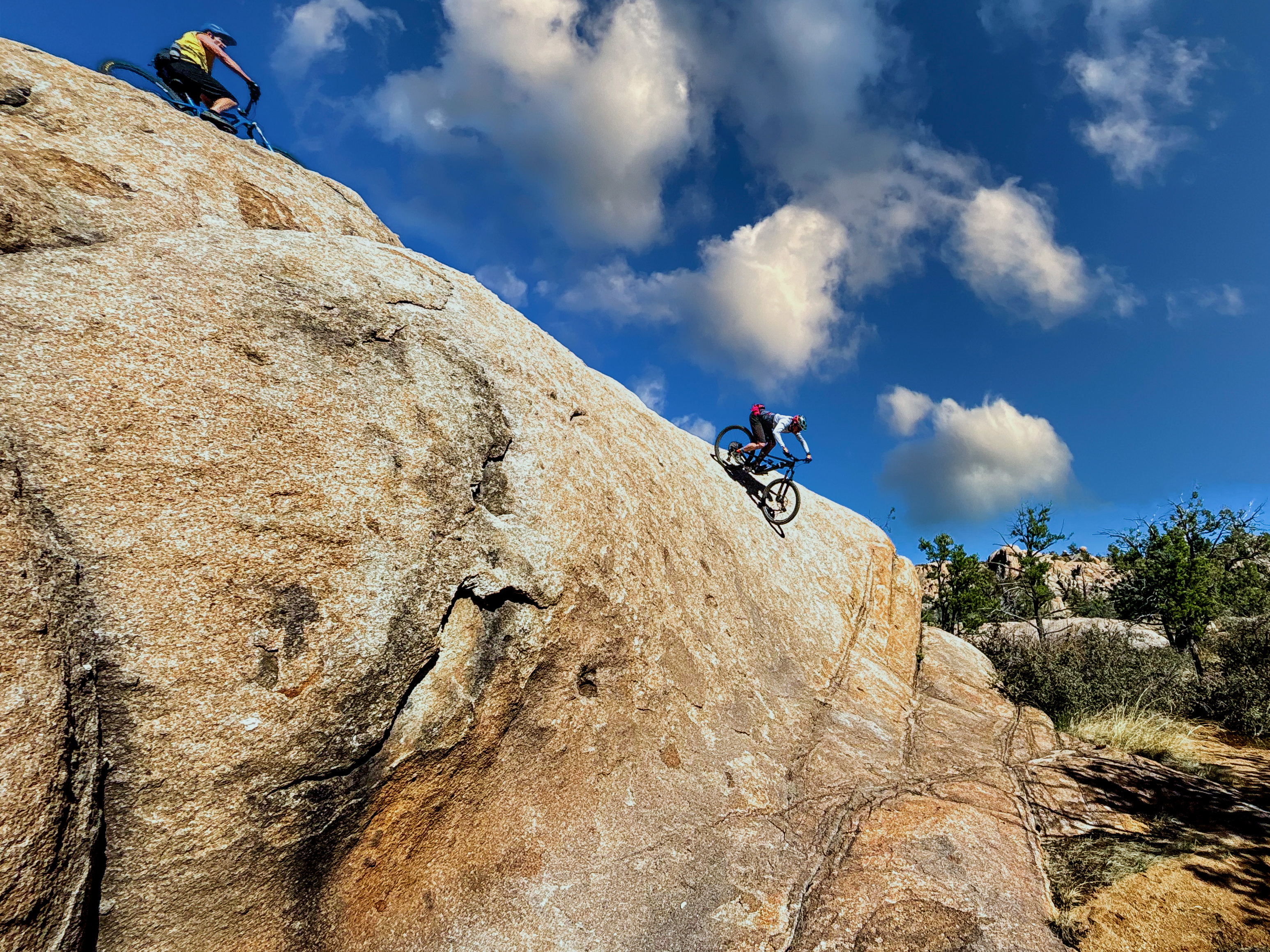 Two mountain bikers descending steep rocky terrain under a bright blue sky with fluffy clouds. The riders navigate large boulders amidst a natural landscape featuring sparse vegetation. Granite Dells mountain bike trail.