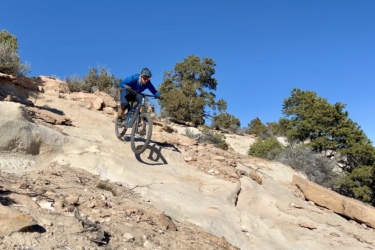 A mountain biker descends a rocky trail under a clear blue sky, surrounded by sparse vegetation and trees. The rider wears a blue long-sleeve shirt and a helmet, focused on navigating the uneven terrain. Spinal Tap mountain bike trail.