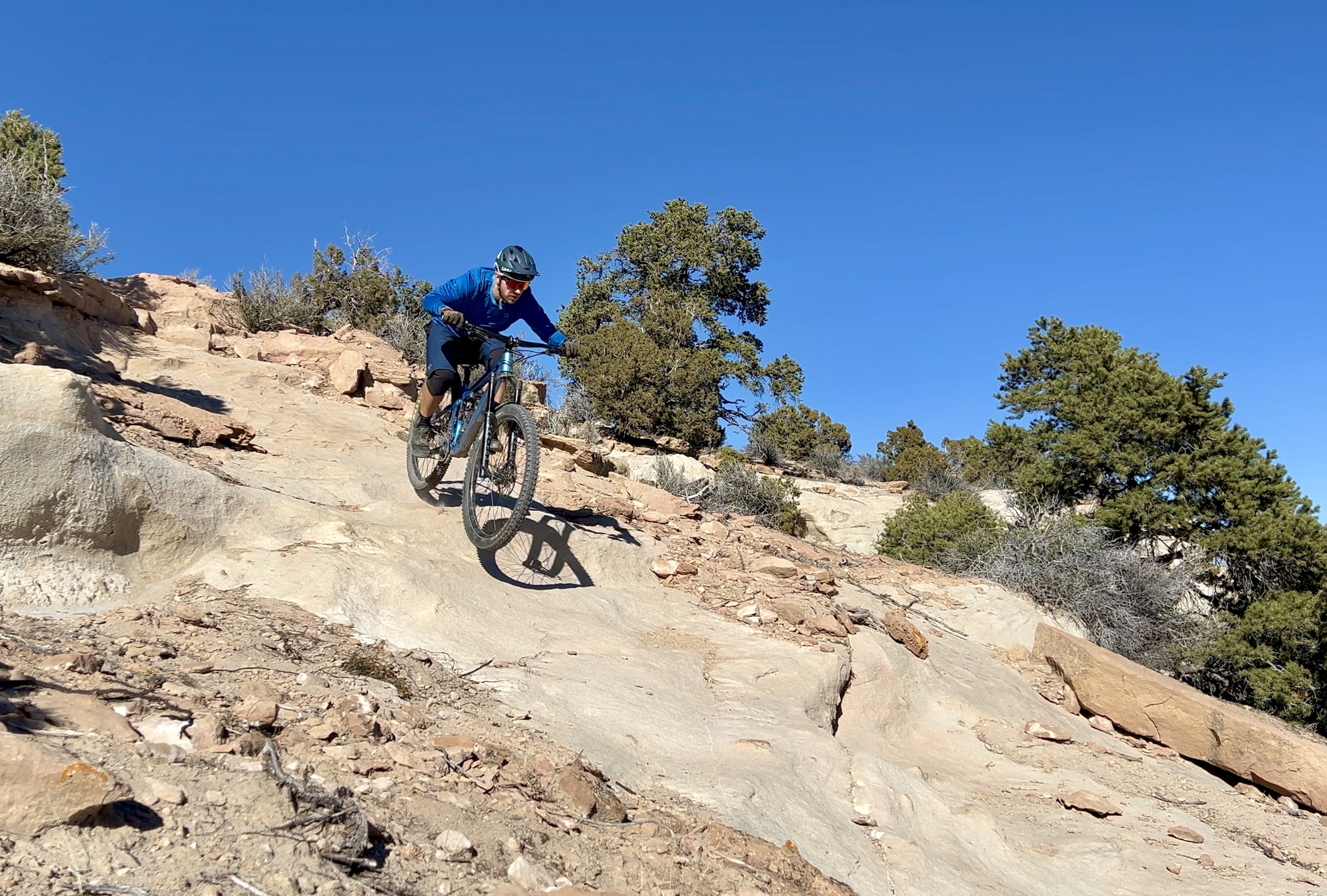 A mountain biker descends a rocky trail under a clear blue sky, surrounded by sparse vegetation and trees. The rider wears a blue long-sleeve shirt and a helmet, focused on navigating the uneven terrain. Spinal Tap mountain bike trail.