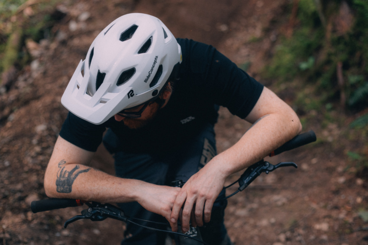 A close-up image of a person wearing a white mountain biking helmet, leaning over their bike's handlebars on a forest trail. The person's arms are tattooed, and they are dressed in a black shirt, with a focus on the bike's controls and the earthen trail in the background.