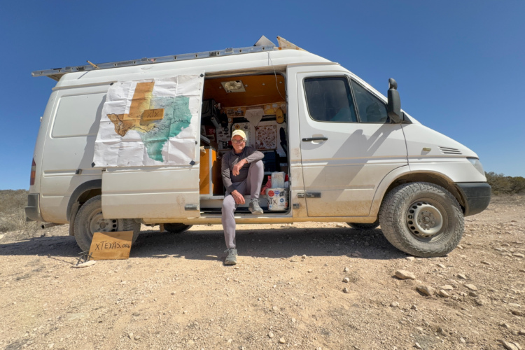 A Van parked in a desert landscape with a Texas map displayed on the side. A person is sitting by the open door, surrounded by various items inside the van. A wooden sign in front reads "xtexas.org." The scene is set under a clear blue sky.
