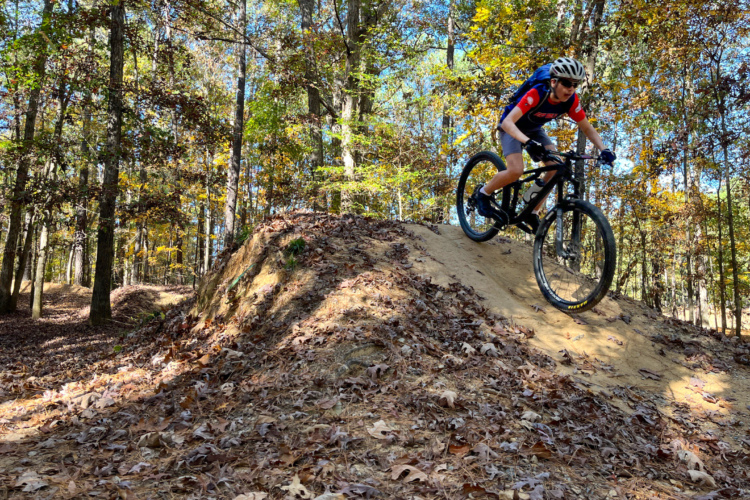 A young rider in a red and black jersey and helmet descends a dirt mound on a mountain bike in a wooded area with autumn foliage.
