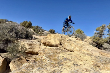 A mountain biker in mid-air, jumping off a rocky ledge, surrounded by sparse vegetation and a clear blue sky. Spinal Tap mountain bike trail.