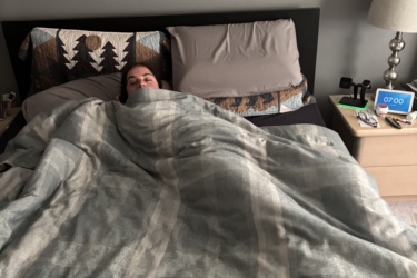 A person is laying in bed under a patterned blanket, with their face barely visible. The bed has a dark headboard and two pillows. In the background, a framed artwork and a bedside table with a lamp and alarm clock are visible. The alarm clock displays the time as 7:00. The Great Trail: Uxbridge mountain bike trail.